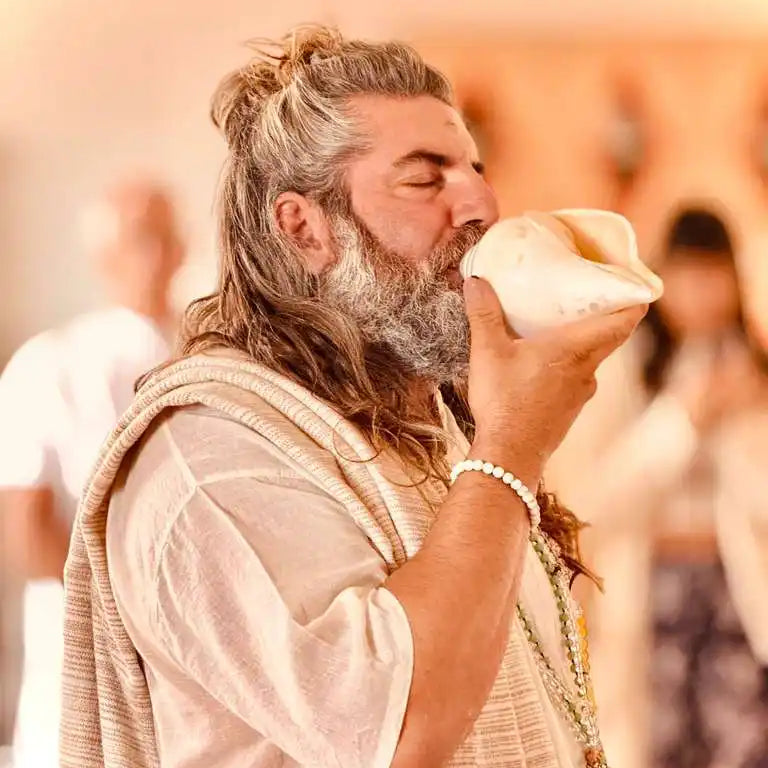 A man with a beard and long hair tied in a bun, wearing traditional attire, blowing into a white conch shell.