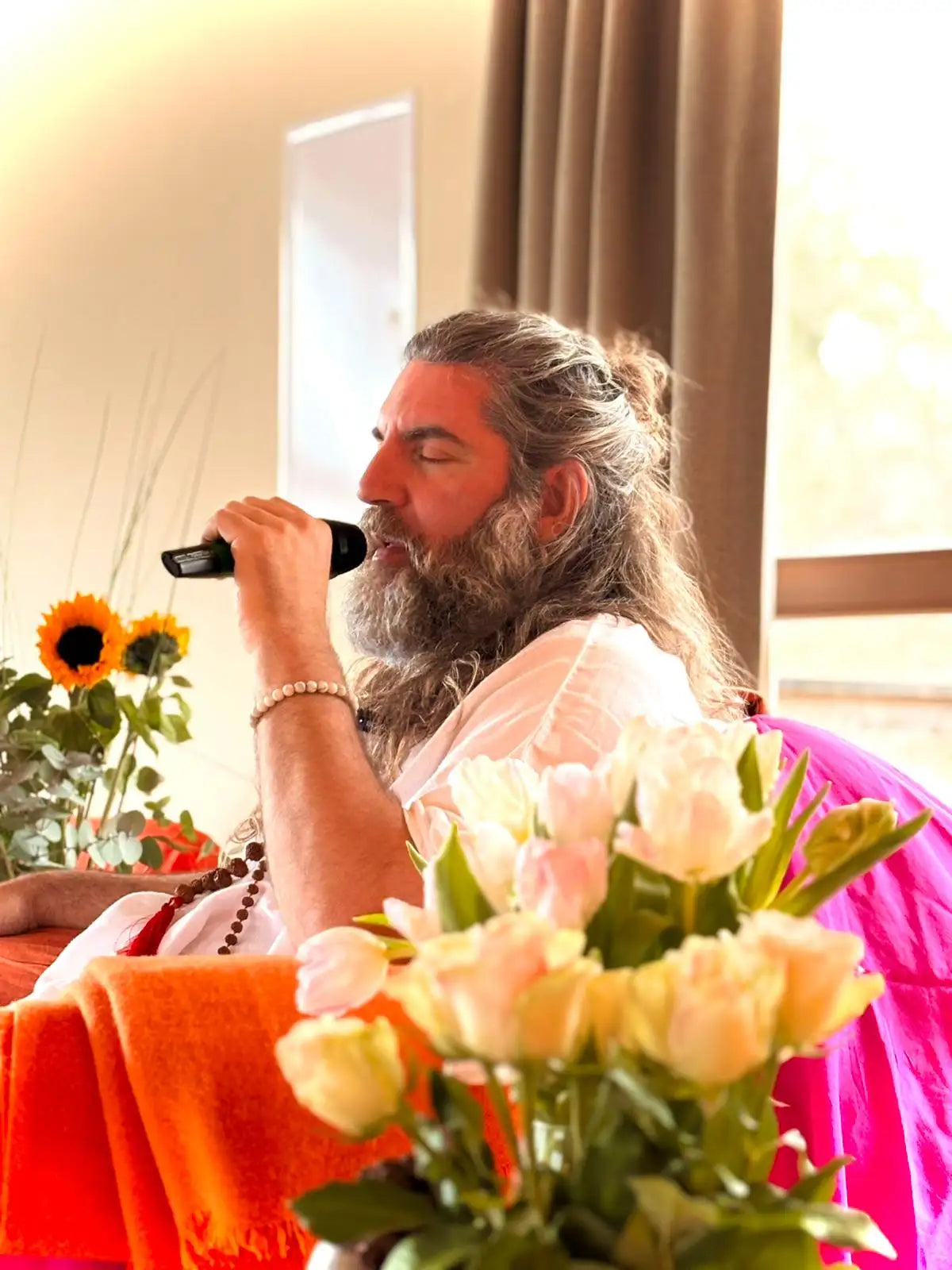 A man with long gray hair and a full beard, wearing a white robe and holding a black microphone.
