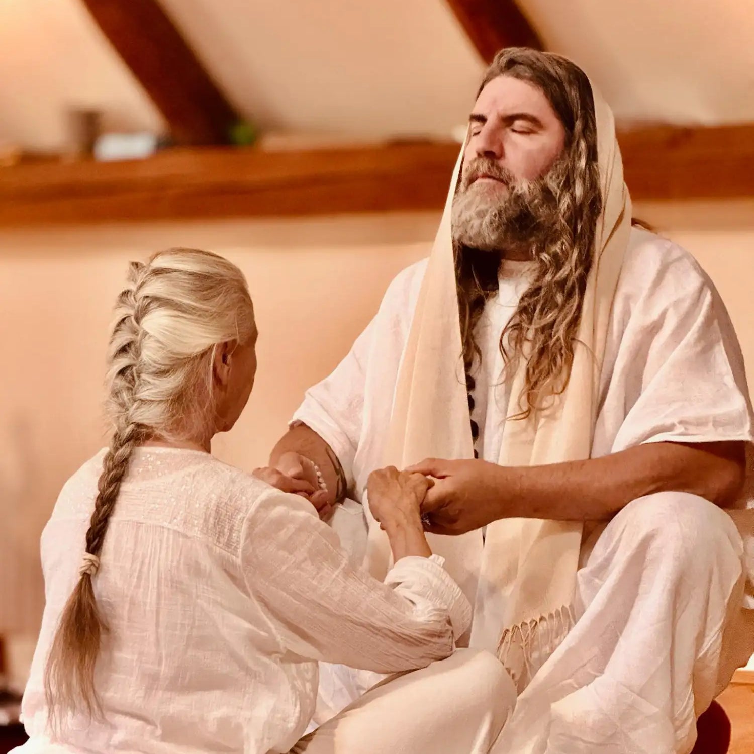 A man with long, wavy brown hair and a full beard wears flowing white robes and a matching draped shawl.