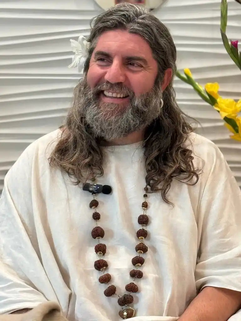 A smiling person with long wavy hair, a full grey beard, and a beaded necklace wearing a white shirt.