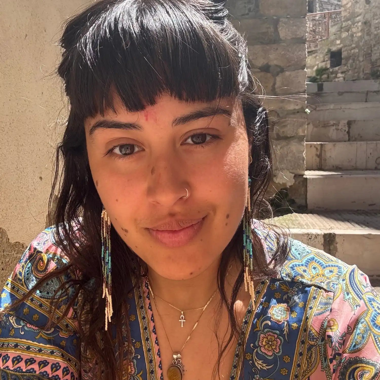 A woman with dark hair and bangs, wearing a patterned blue and pink top, long beaded earrings, and several necklaces.